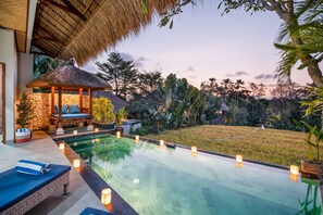 A swimming pool with candles, a thatched-roof gazebo, and a stone wall. - Villa Anyar (Canggu)