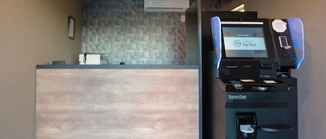 Un mostrador de recepción con una pantalla digital que muestra información de habitaciones, un quiosco de autoservicio SystemGear y una pared con azulejos al fondo.