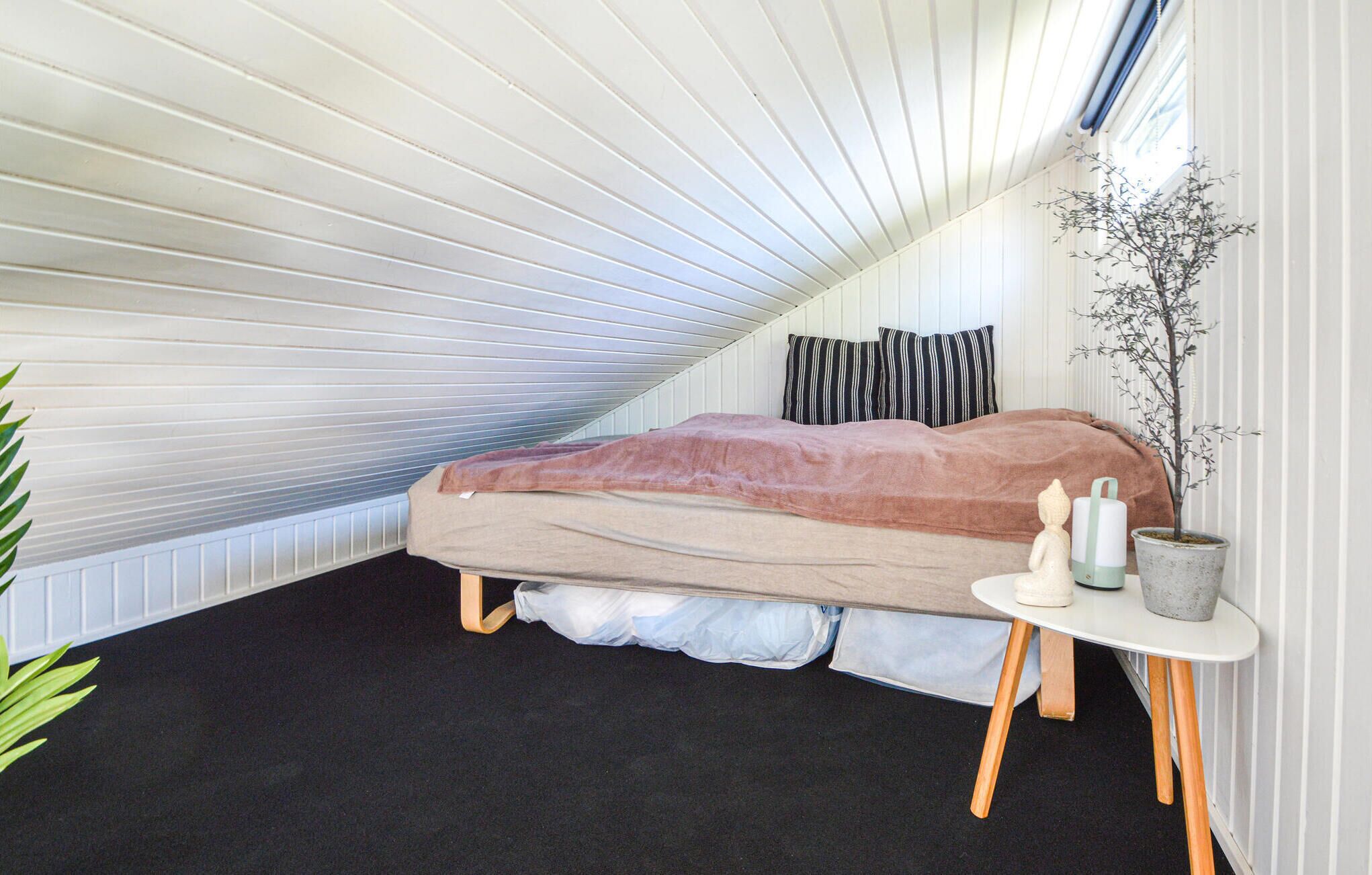 A bedroom with a sloped ceiling, a bed with a pink blanket, a small bedside table with candles, and a potted plant.