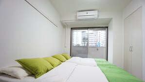 Ein Schlafzimmer mit Bett, grünen Kissen, einer weißen Bettdecke, einem Fenster mit Blick auf Gebäude und einer Klimaanlage.