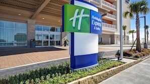 A Holiday Inn Express building with a prominent sign, large windows, and a landscaped area with plants and a palm tree. - Holiday Inn Express And Suites Galveston Beach (Galveston)