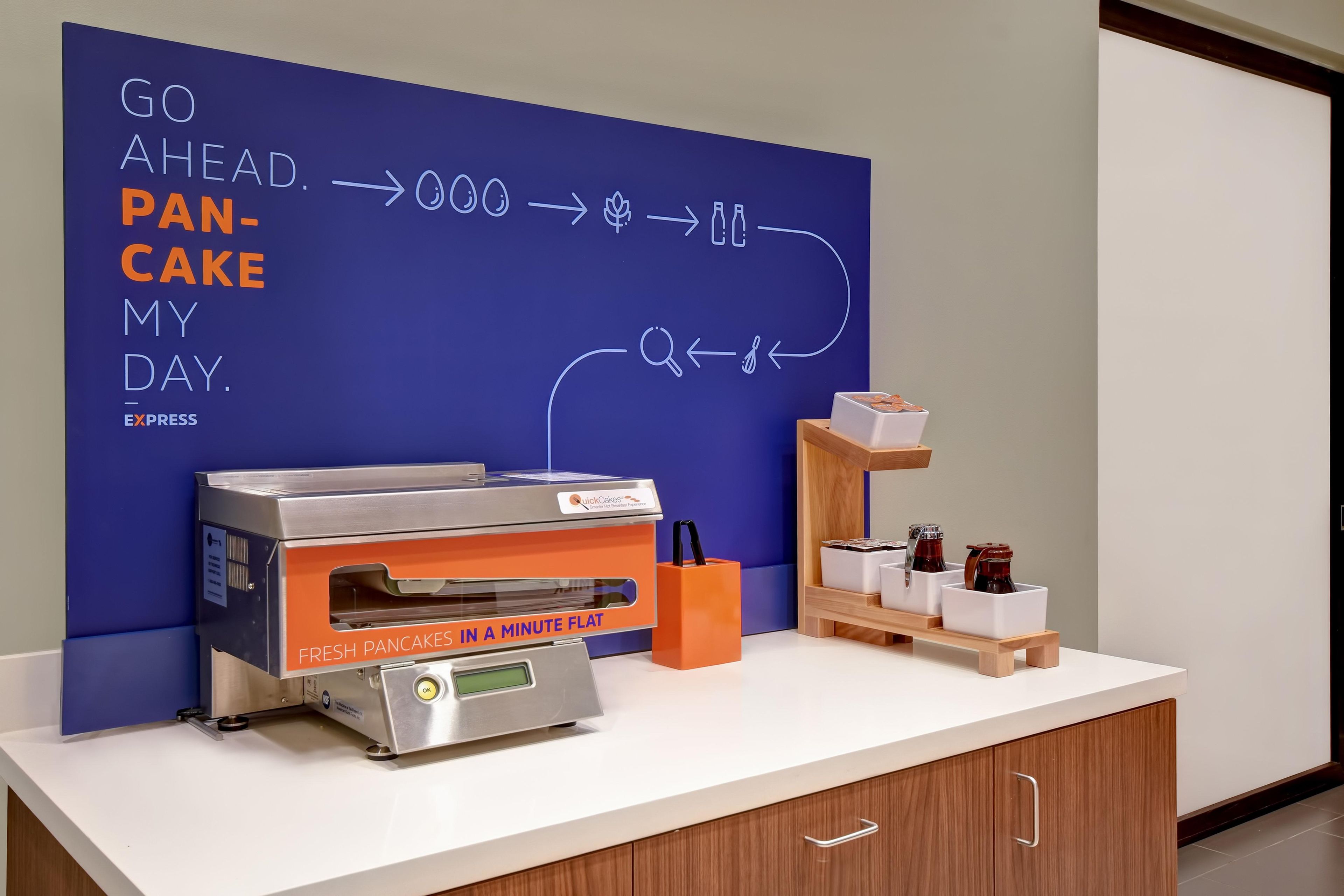 Un plan de travail de cuisine avec une machine à crêpes QuickCake Express, un mur bleu orné d’inscriptions motivantes et une étagère en bois contenant des ustensiles de cuisine.