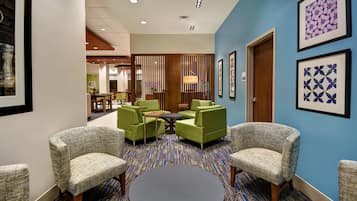 A hotel lobby with a patterned carpet, green armchairs, a round table, and framed artwork on the walls.