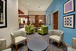 Un hall d’hôtel avec un tapis à motifs, des fauteuils verts, une table ronde et des œuvres d’art encadrées accrochées aux murs.