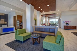 Eine moderne Lobby mit grünen gepolsterten Sofas, einem Empfangstresen, einem mit Snacks gefüllten Kühlschrank und einer Sitzecke mit einem gemusterten Sofa und einem kleinen Tisch.