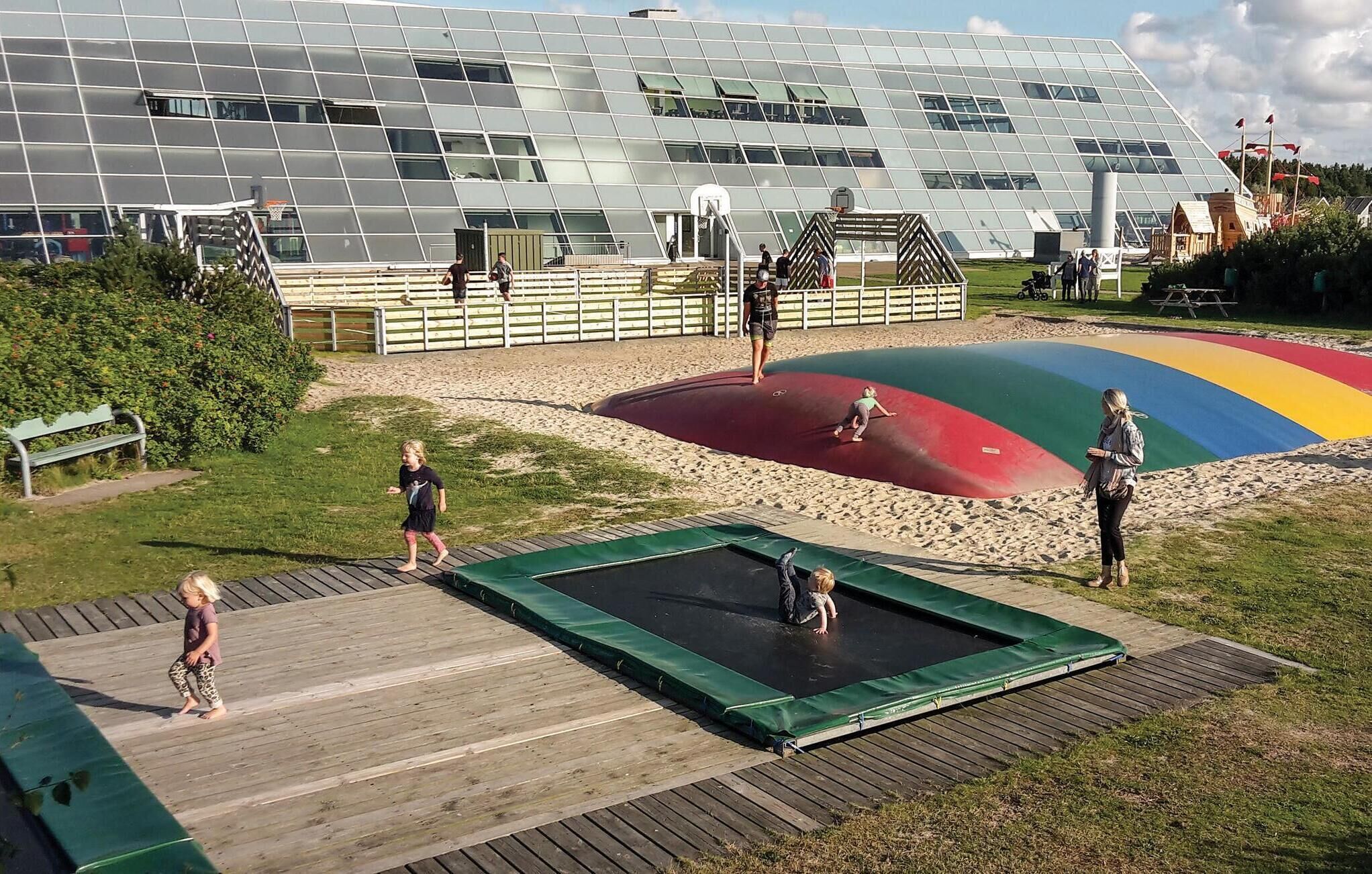 Un parque infantil con un trampolín y un tobogán colorido, rodeado de una terraza de madera y césped.