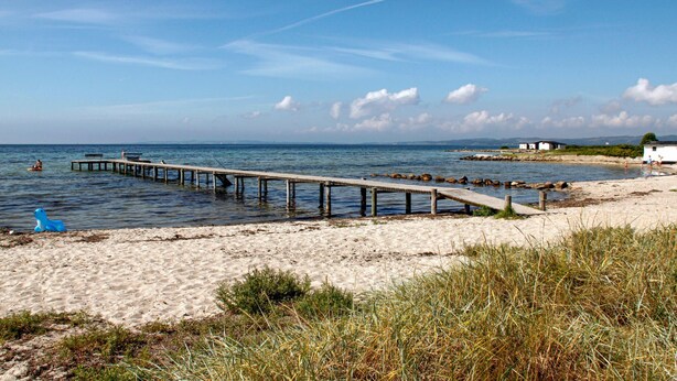 Une plage avec une jetée en bois qui s’avance dans l’eau, un rivage sablonneux et un petit bâtiment en arrière-plan.