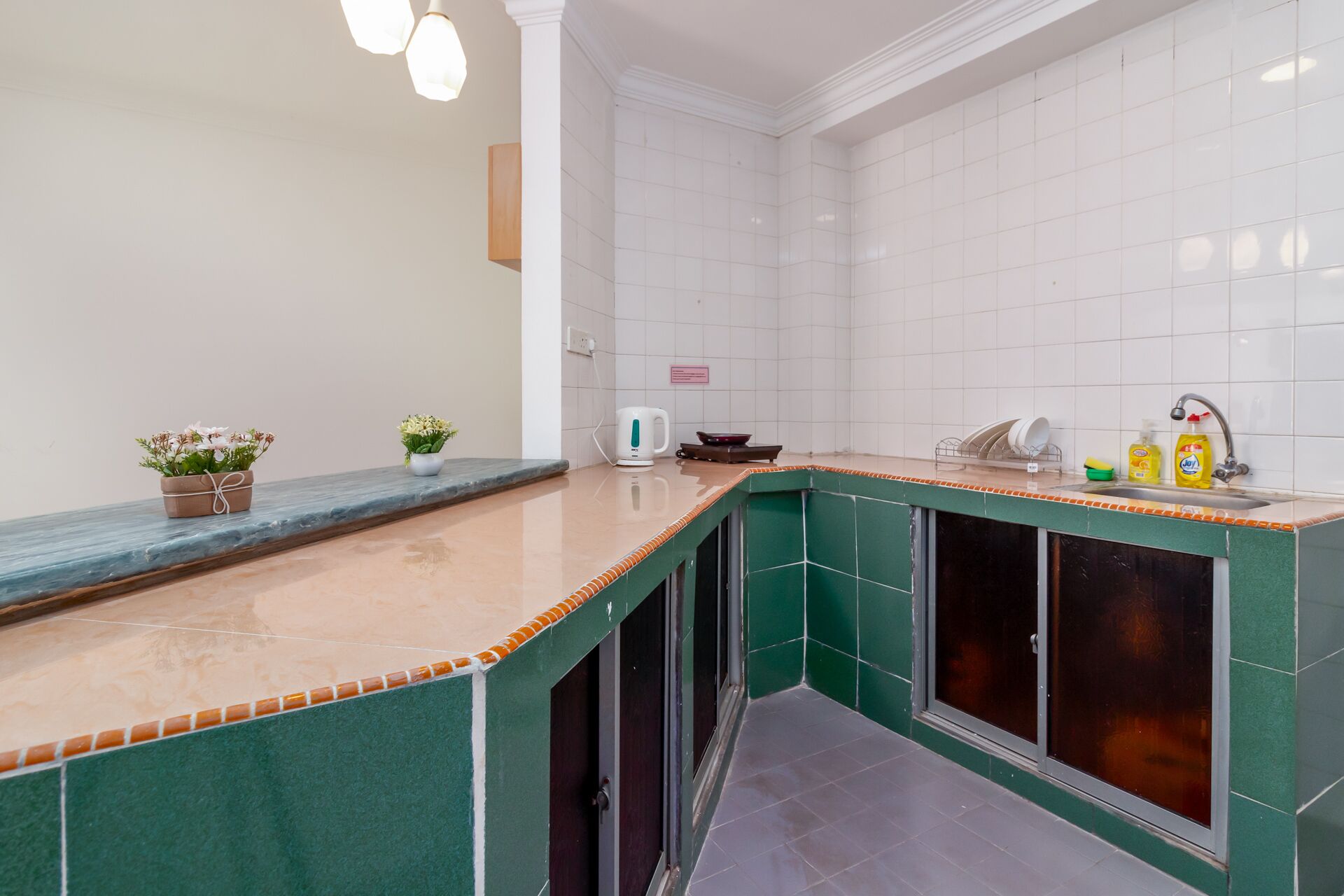 A kitchen with a green countertop, white tiled walls, and a sink with dish soap and a kettle.