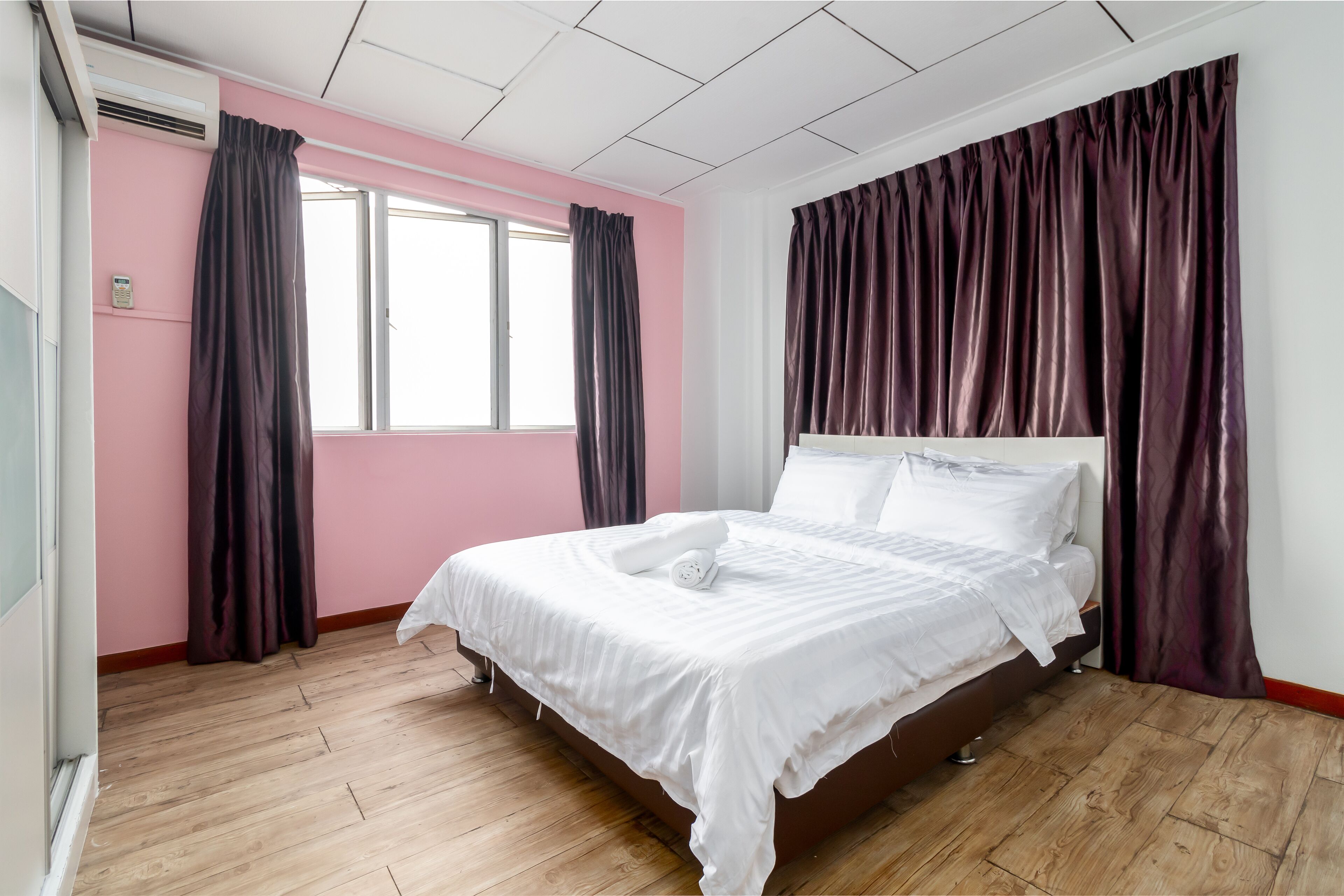 A bedroom with a wooden floor, a bed with white bedding, pink walls, and dark curtains.