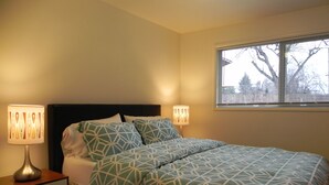 A neatly made bed with patterned bedding, two bedside lamps, a nightstand, and a window with a view of trees. - Mid-Century Modern w/ Garage Close to Amenities & 3 Miles to Autzen/UO! (Eugene)