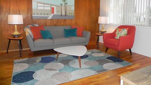 A living room with a grey sofa, red armchair, and a coffee table. There are two lamps on side tables, a colorful rug, and a painting on the wall. - Mid-Century Modern w/ Garage Close to Amenities & 3 Miles to Autzen/UO! (Eugene)