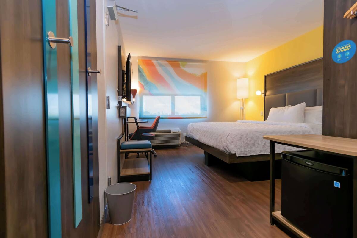 A hotel room with a bed, a desk, a chair, a trash bin, and a wall with a colorful abstract design.