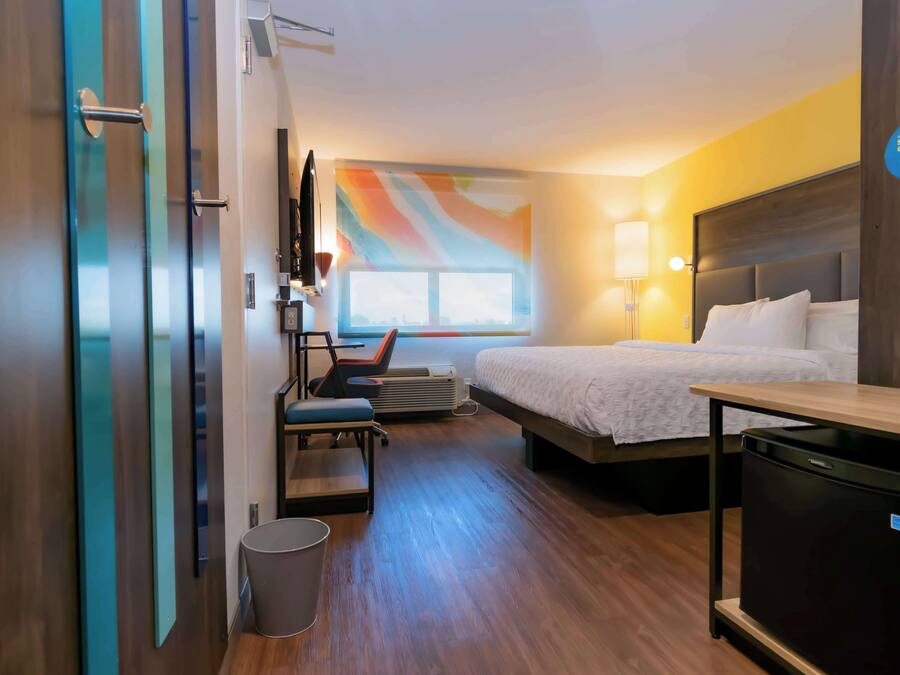 Une chambre d’hôtel comprenant un lit, un bureau, une chaise, une poubelle et un mur orné d’un motif abstrait aux couleurs vives.