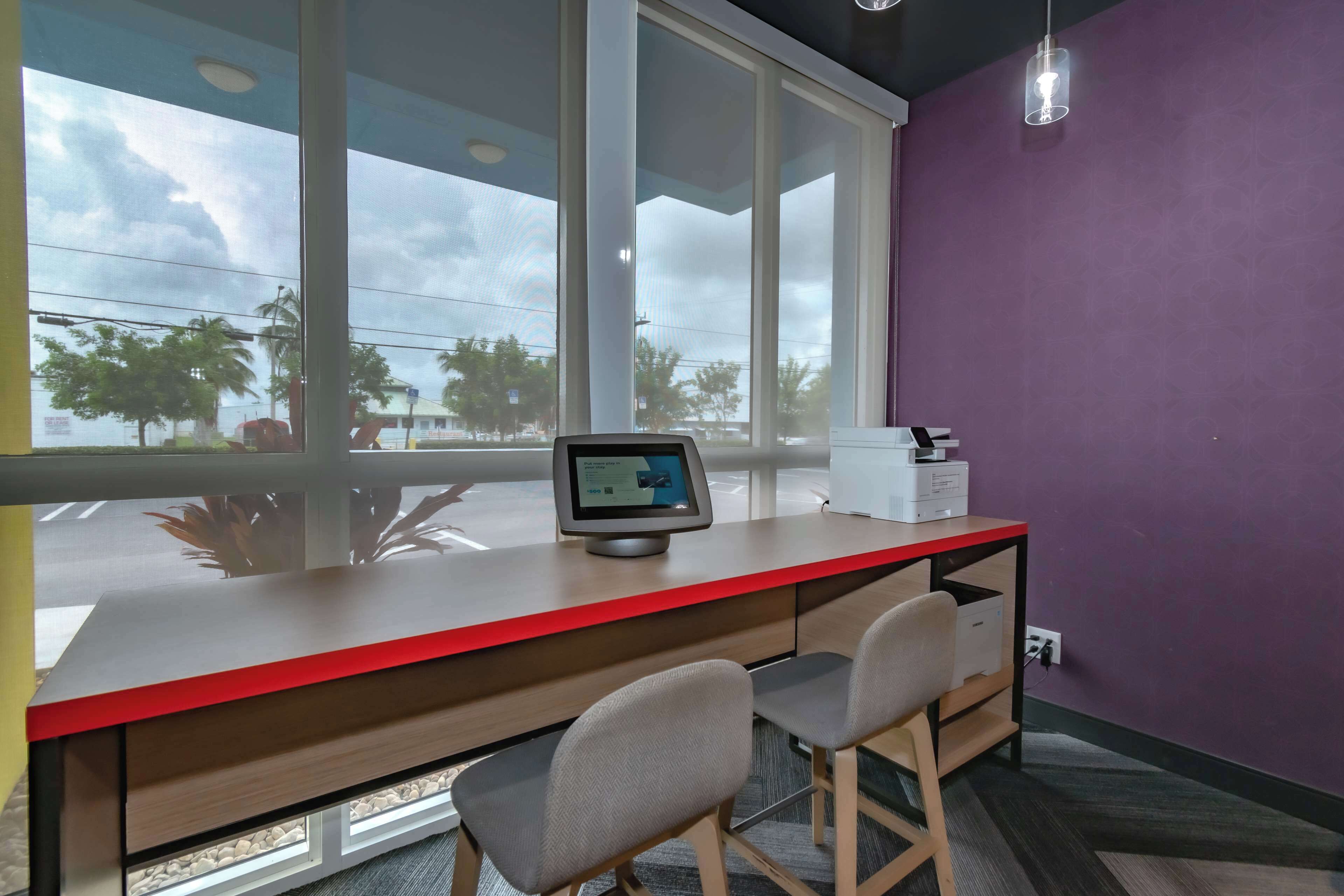 A modern office space with a desk, chairs, a computer monitor, and a window with a view of palm trees and buildings.