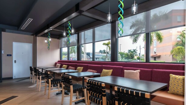 Een moderne eetruimte met houten tafels en stoelen, een rode, beklede bank en grote ramen met uitzicht op palmbomen en gebouwen.