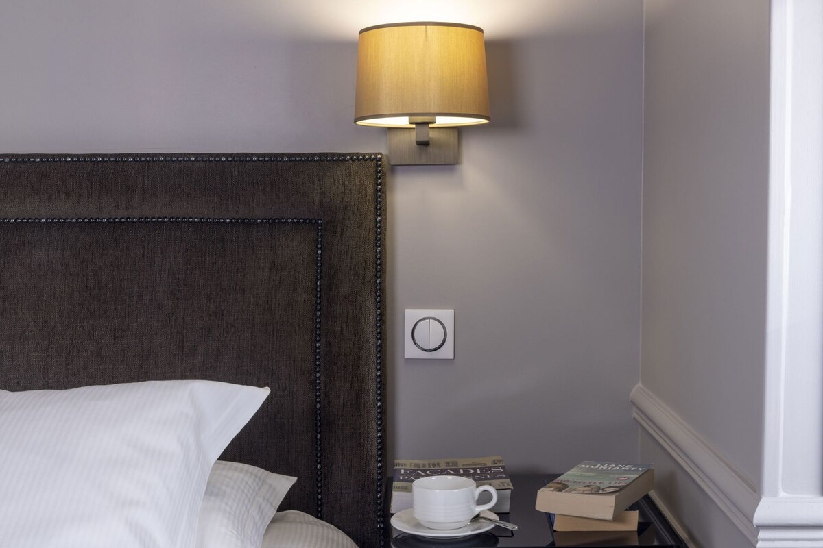 A hotel room with a bed, a nightstand with a lamp, a cup, and books.