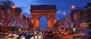 Una calle concurrida con una estructura de arco destacada, probablemente un monumento histórico, vehículos y árboles iluminados.