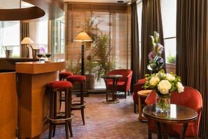 Un vestíbulo de hotel con una recepción, sillas rojas, una mesa redonda con un jarrón de flores y un ventanal con persianas.