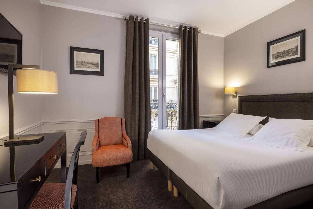Une chambre d’hôtel avec un lit, un bureau, une chaise, une lampe et une fenêtre avec des rideaux.