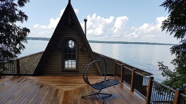 A wooden deck with a hammock chair, a wooden cabin with a triangular roof, and a view of the water. - BEAUTIFUL FOUR SEASON WATERFRONT COTTAGE (Innisfill)
