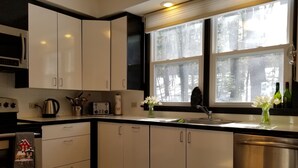 A modern kitchen with white cabinets, stainless steel appliances, and a large window with a view of trees. - BEAUTIFUL FOUR SEASON WATERFRONT COTTAGE (Innisfill)