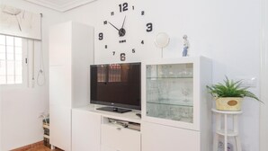 En stue med et fladskærms-tv på et hvidt skab, et ur over skabet, og en glasmontre med vinglas.