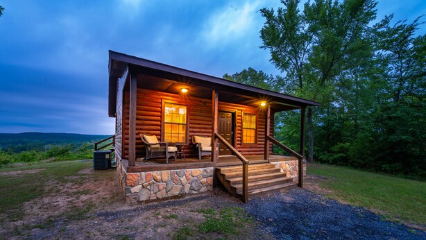A log cabin with a porch, stone foundation, and a view of the surrounding landscape. - The Ossi is a secluded couples cabin with panoramic mountain and sunset views! (Smithville)