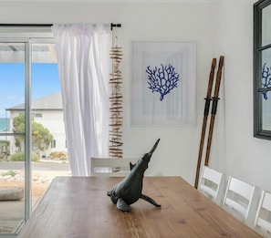 A dining area with a wooden table, white chairs, a glass door leading to a view of the ocean, and a wall with a coral artwork.