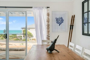 A dining area with a wooden table, white chairs, a decorative wall hanging, and a view of the ocean through a glass door.
