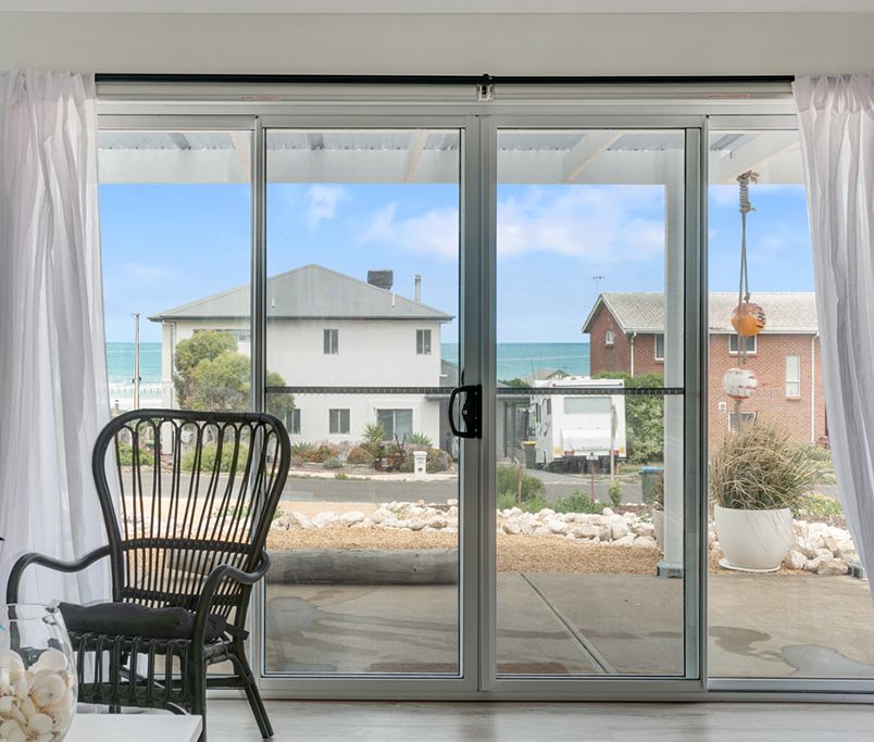 A room with two chairs, a table, and a view of the sea through a sliding glass door.