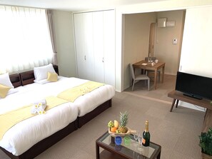 Quarto de hotel com cama, escrivaninha, televisão e mesa de centro com fruta e uma garrafa.
