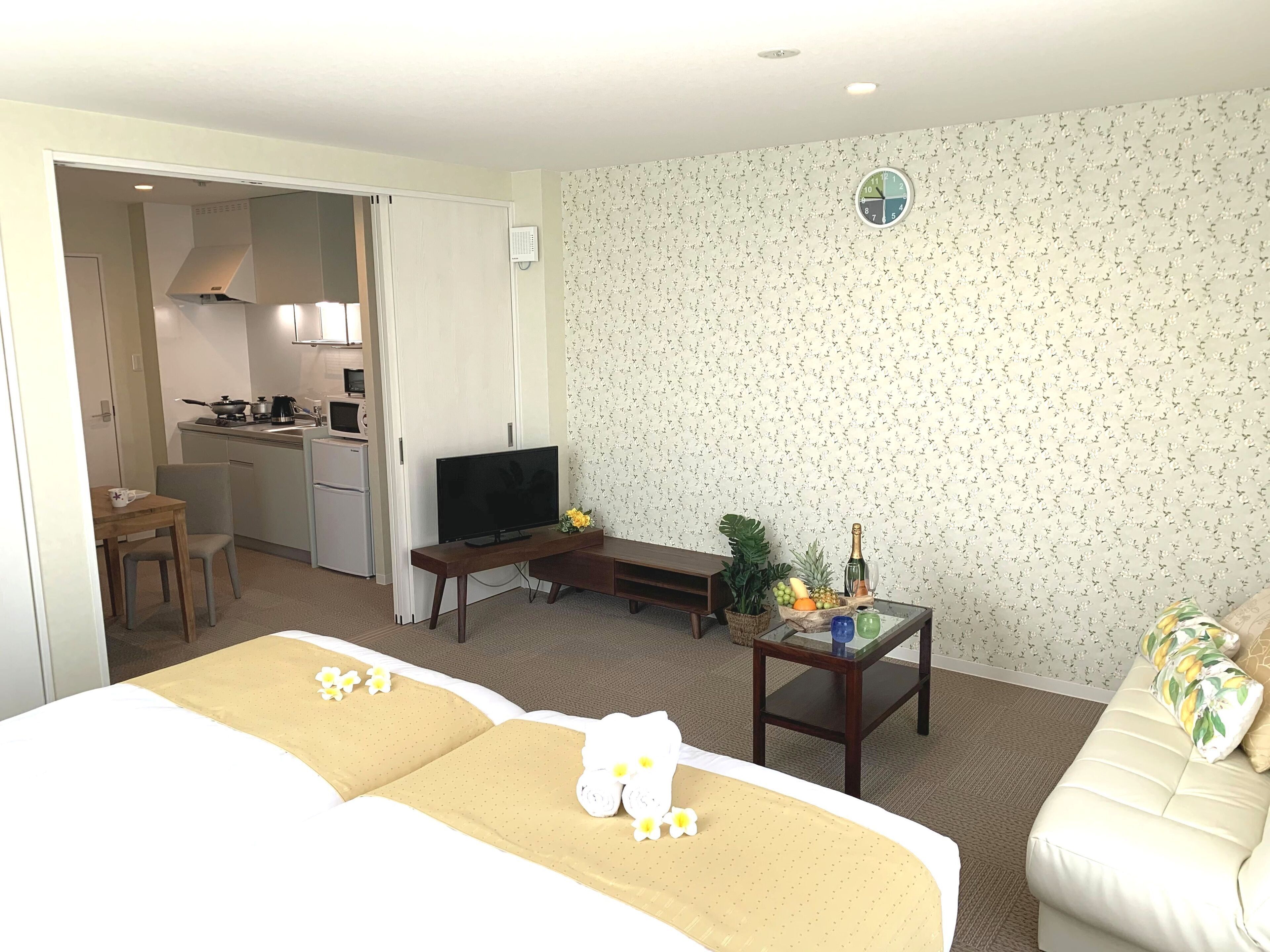 A hotel room with a bed, a TV, a small desk with a plant, and a kitchenette in the background.
