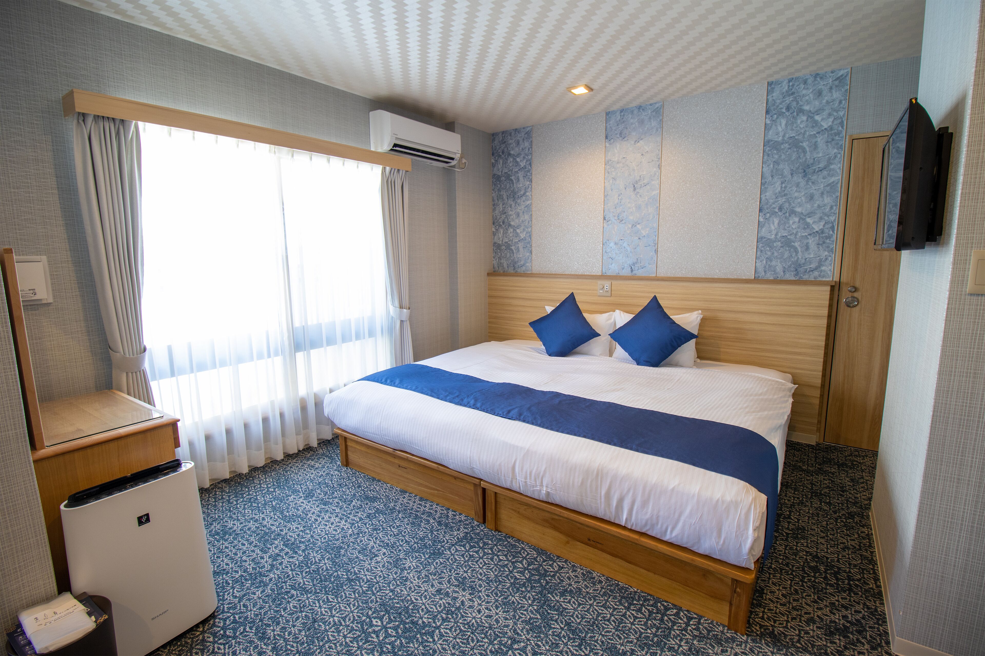 Une chambre d’hôtel avec un grand lit, une tête de lit en bois, un téléviseur à écran plat fixé au mur et une fenêtre avec des rideaux.