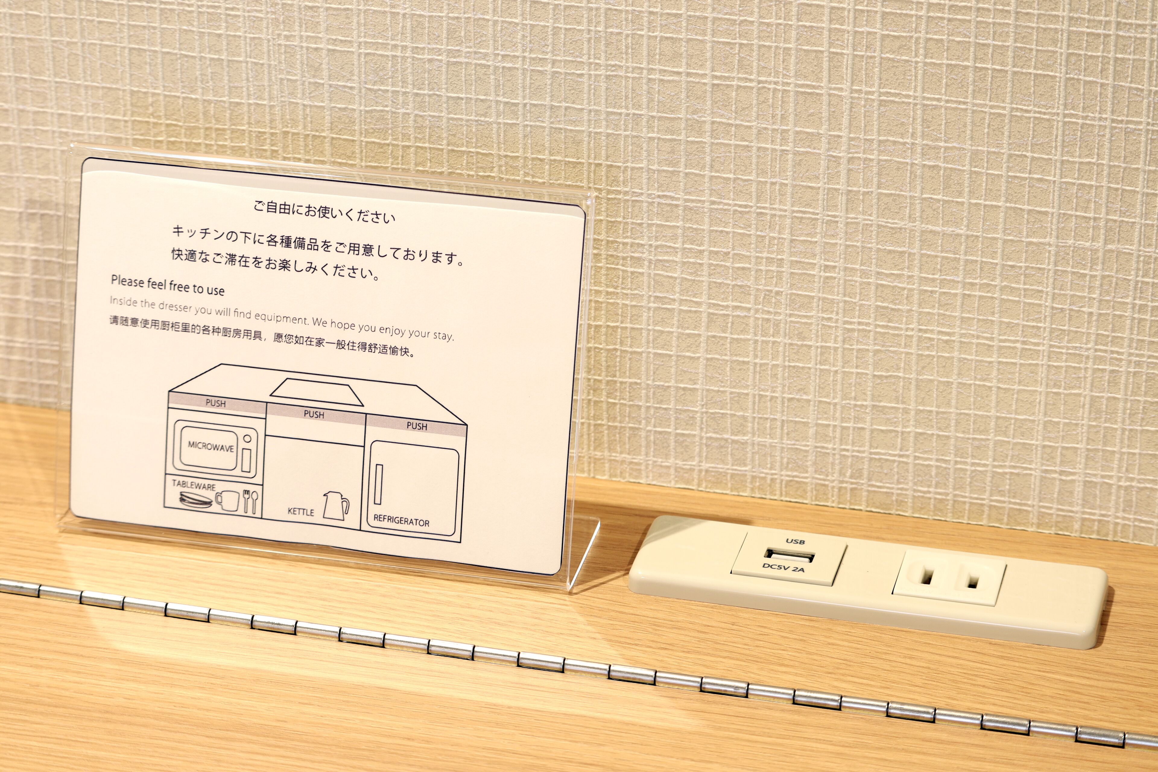 A hotel room with a kitchenette area, including a microwave, table, kettle, and refrigerator. There is a power strip with a USB port and a power outlet.