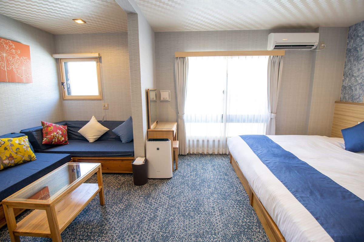 Une chambre d’hôtel comprenant un lit, un canapé, une petite table, un bureau, un appareil de climatisation et une fenêtre avec des rideaux.