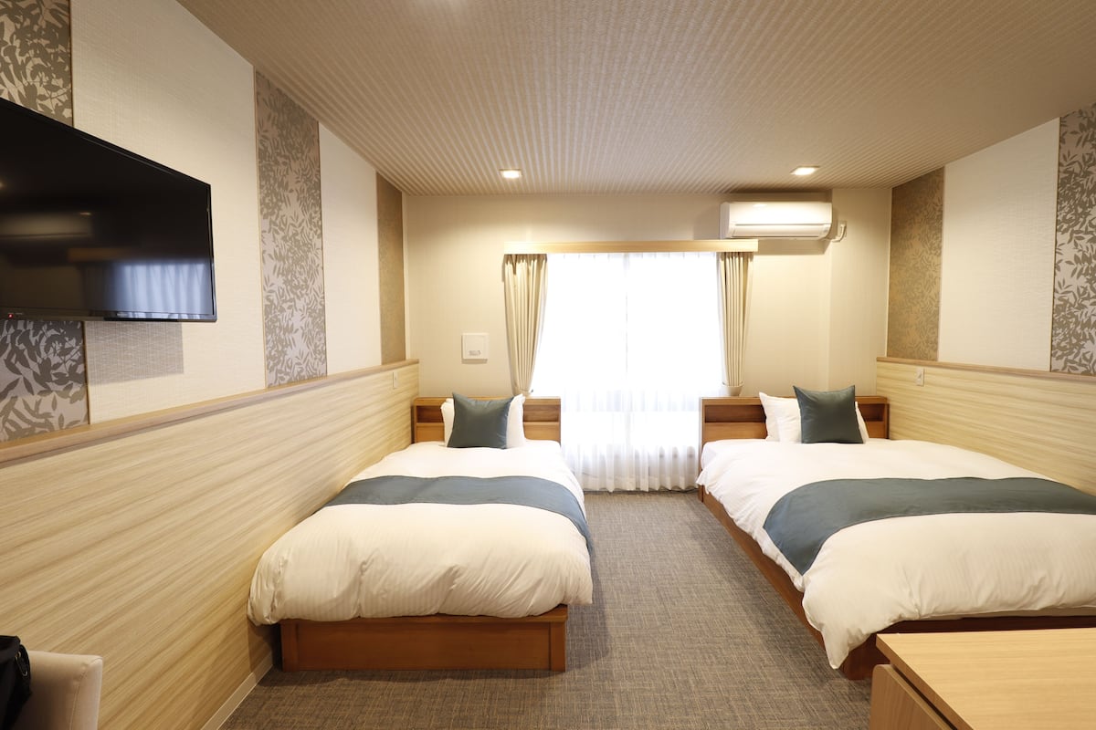 A hotel room with two beds, a flat-screen TV, an air conditioning unit, and a window with curtains.