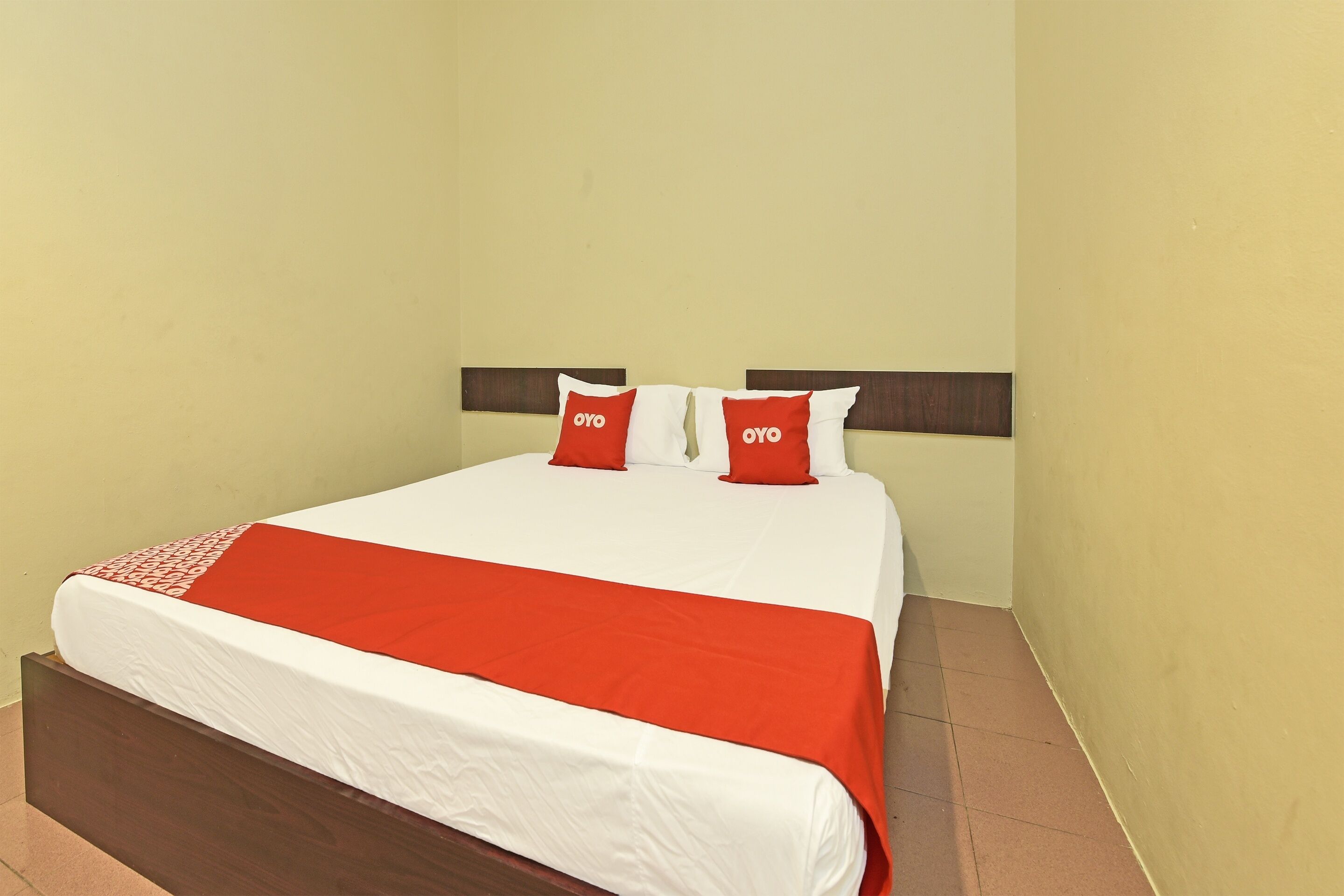 Una cama bien tendida con una colcha blanca y roja, dos almohadas rojas con la marca 'OYO', y un cabecero de madera.