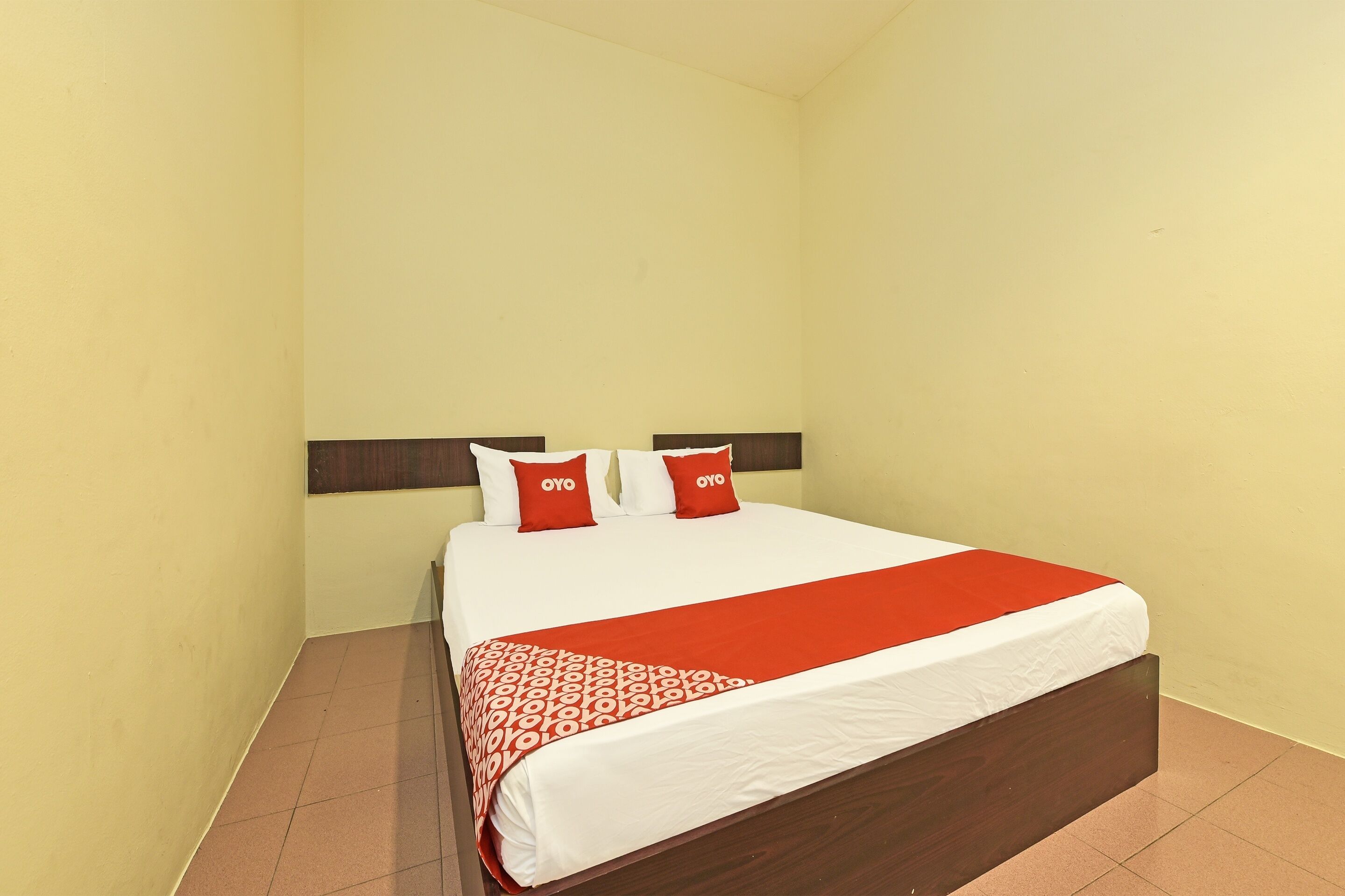 Habitación de hotel con una cama grande, dos almohadas rojas, una colcha blanca y roja, y un cabecero de madera.