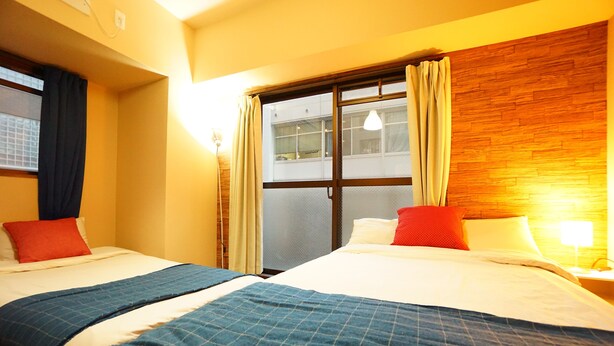 A hotel room with two beds, a wooden headboard, a window with curtains, and a nightstand with a lamp. - Terry's Apartment Shinsaibashi East I G04C (Osaka)