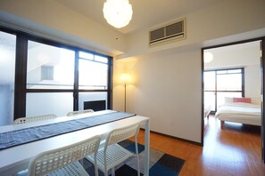Una habitación con una mesa blanca, dos sillas blancas, una lámpara y una cama con un almohá de color rojo.