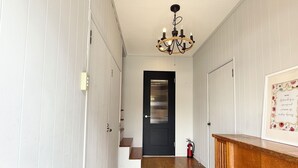 A hallway with white paneled walls, a wooden floor, a black door, and a chandelier. - Tabisai House (Fukuoka)