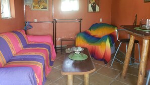 A room with two beds, a table, and a chair. There are paintings on the walls and a window with curtains.