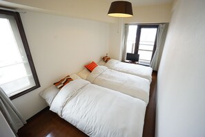 Two single beds with white bedding and a brown headboard, a table with a lamp and TV, and a large window with curtains.