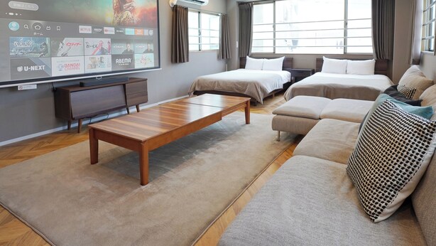 Une chambre d’hôtel moderne dotée d’un grand écran de télévision diffusant divers services de streaming, d’une table basse en bois, de deux lits, d’un canapé et d’une grande fenêtre laissant entrer la lumière naturelle.