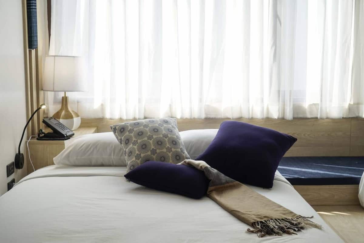 Una habitación de hotel con cama, lámpara de noche, teléfono y ventana con cortinas traslúcidas.