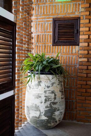 Un grand bac à fleurs, usé par les intempéries, contient des plantes vertes devant un mur de briques orné d’une fenêtre et d’une porte.