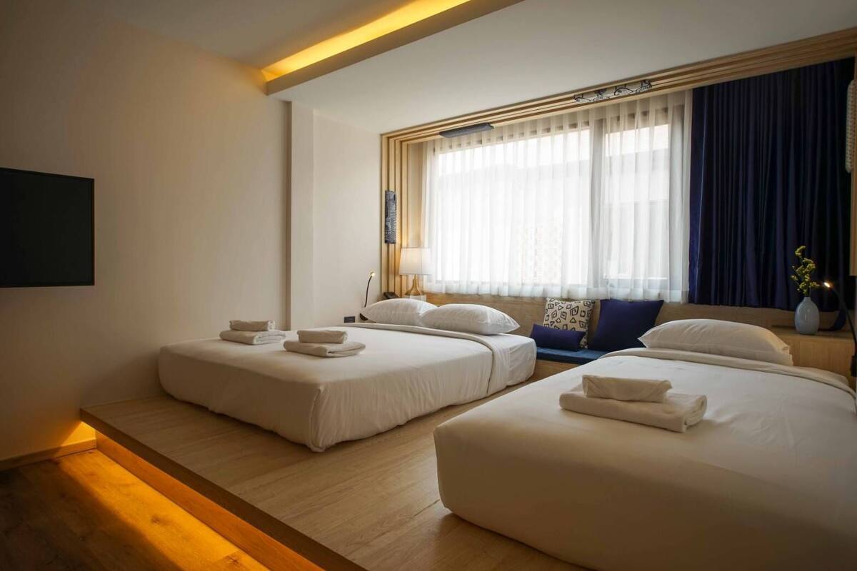 Habitación de hotel con dos camas, una mesita de noche, una lámpara y un ventanal con cortinas.