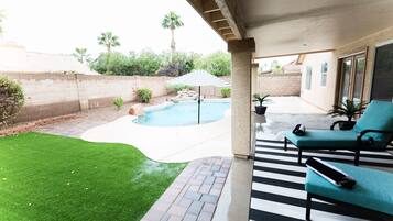 A backyard with a swimming pool, a patio area with a striped umbrella, and a seating area with two lounge chairs.