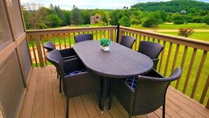 Una terraza de madera con una mesa redonda y seis sillas de mimbre, con vistas a un paisaje verde con árboles y una casa distante.