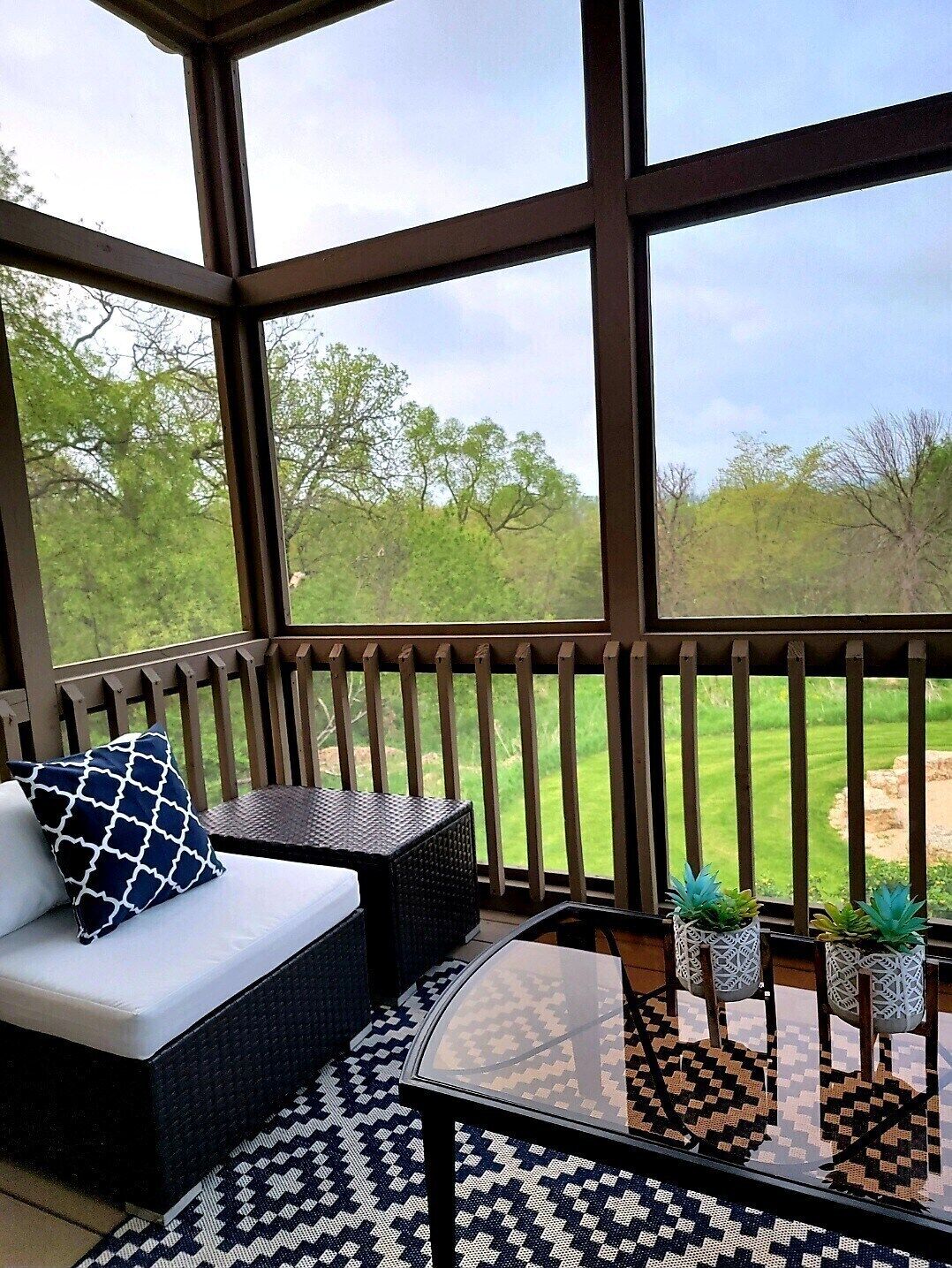 A covered outdoor seating area with a sofa, cushions, and a glass table with potted plants.